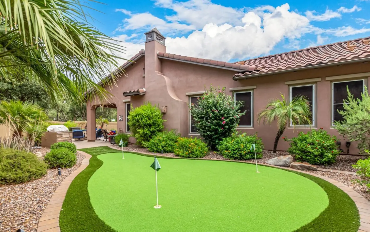 A Scottsdale backyard features a small artificial putting green surrounded by rocks and lush greenery. The house boasts a stucco exterior with a red-tiled roof, while a palm tree and various shrubs adorn the landscape under a partly cloudy sky.