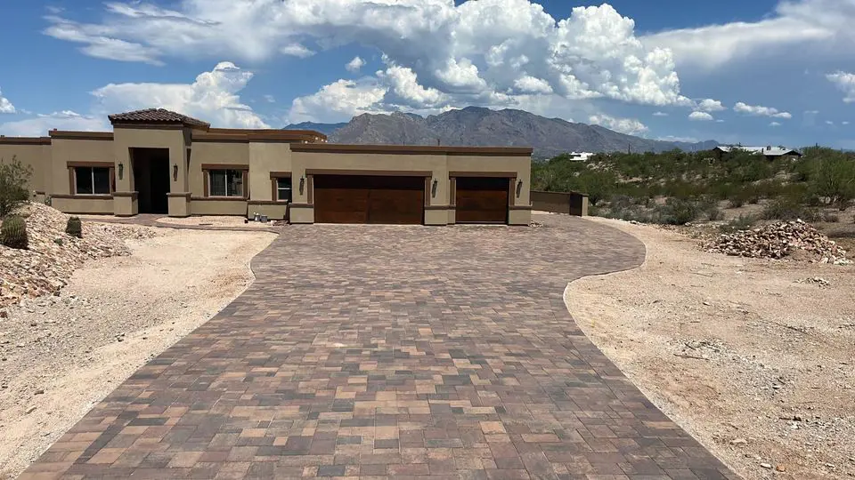 A modern, single-story house with a flat roof is surrounded by desert landscaping in Scottsdale, AZ. The long driveway, paved with travertine paver, leads to two wooden garage doors. Mountains and a partly cloudy sky provide a stunning natural backdrop.