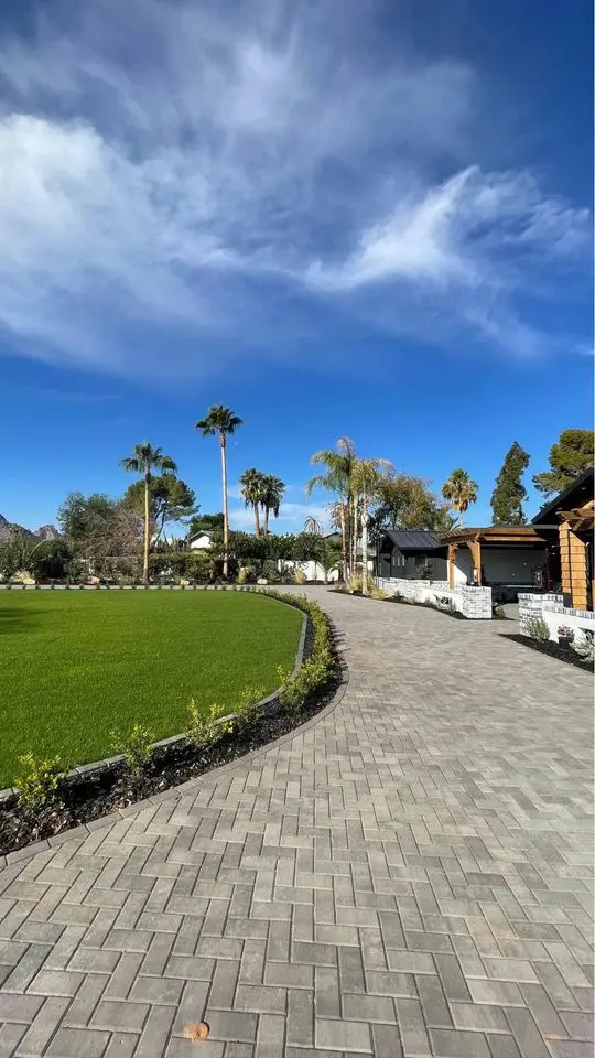 A paved pathway, crafted from elegant travertine pavers, curves through a green lawn bordered by shrubbery, leading to a building. Palm trees stand tall under Scottsdale AZ's vivid blue sky with wispy clouds.