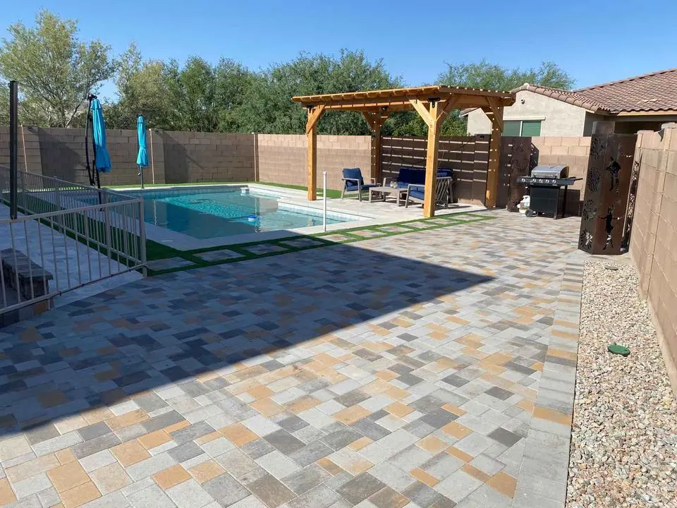 The backyard features a pool encircled by a stylish fence, stone patio, and a pergola with cozy seating. Umbrellas and a grill enhance the space while lush trees and meticulous gravel landscaping in Scottsdale, AZ, complete the scene under clear skies.