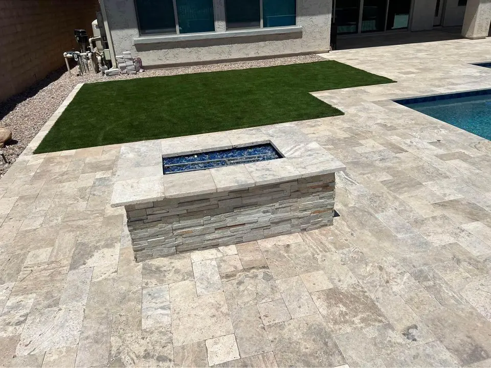 A stone patio featuring a rectangular fire pit with dark stones at its center enhances the backyard setting. Surrounded by natural stone tiles and adjacent to a pool, it showcases a blend of textures, including a patch of artificial grass. This elegant design is reminiscent of Scottsdale, AZ aesthetics.