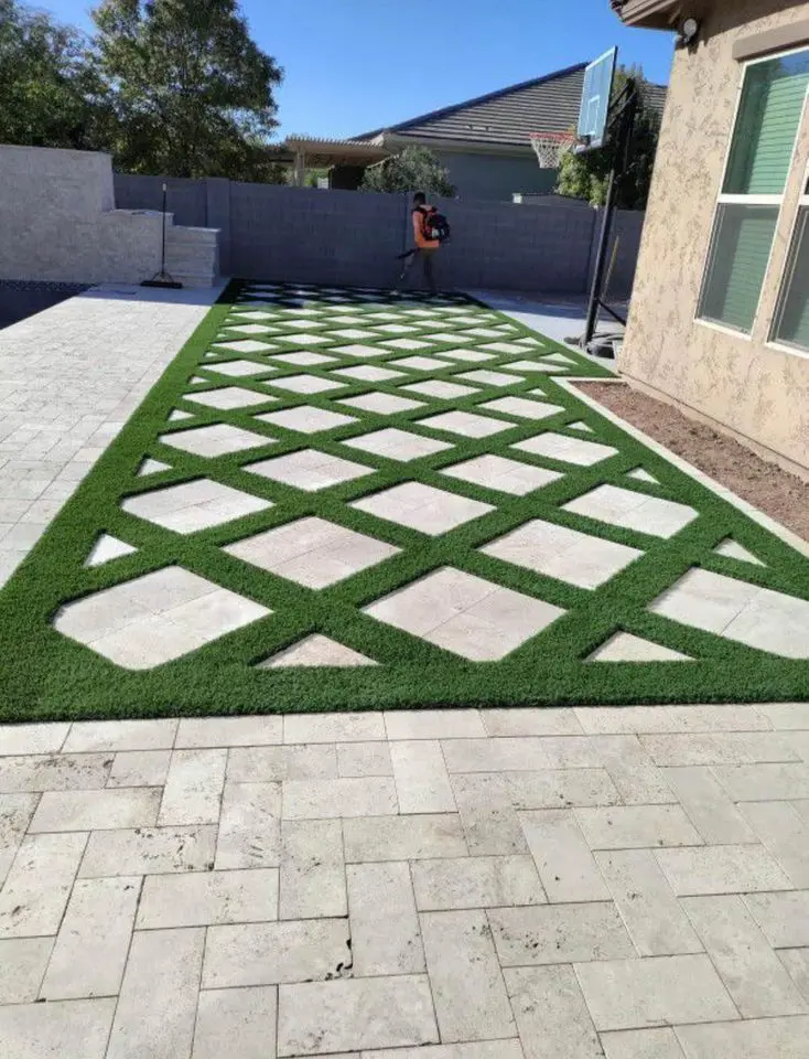 A backyard with a modern geometric design features perpendicular pathways of grass and travertine pavers. A person in the background is using a leaf blower. There is a basketball hoop on the right and a sunny, clear Scottsdale AZ sky above.