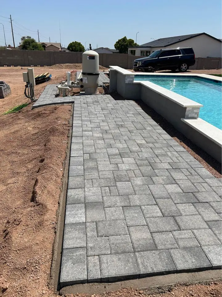 A backyard scene showcases a partially finished travertine paver walkway leading to a swimming pool. Construction materials and equipment are visible nearby, hinting at ongoing work. A black SUV is parked in the background under Scottsdale AZ's clear and sunny sky.