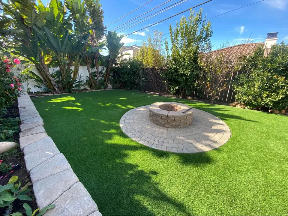 A Scottsdale backyard showcases a winding brick path flanked by expertly installed artificial grass and gravel. A palm tree stands gracefully near the brick wall, while a covered patio invites relaxation on the right.