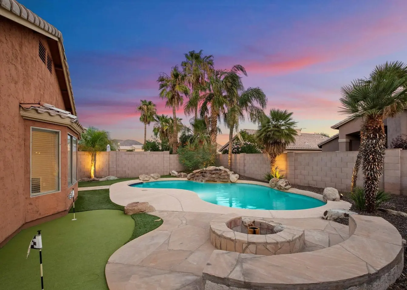 A Scottsdale backyard featuring a swimming pool, fire pit, and putting green. Palm trees and a stone wall surround the area with lush artificial grass installed by expert turf installers. The sky displays vibrant hues of pink and purple at sunset, creating a stunning desert oasis.