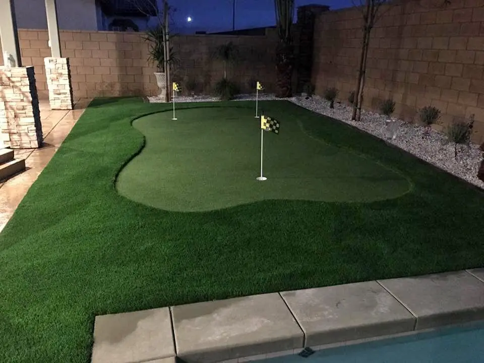 A backyard golf putting green in Scottsdale, featuring artificial grass installation with two flags on a neatly trimmed lawn, is bordered by a stone wall and garden beds, all beautifully illuminated in the evening.