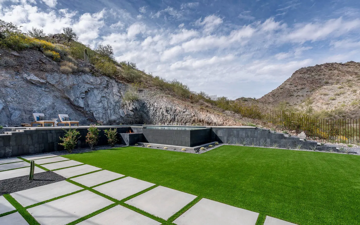 A modern backyard in Scottsdale showcases a green artificial lawn expertly crafted by top artificial turf installers. A sleek, low-profile black stone water feature enhances the space, flanked by rocky hills and blue skies. Two lounge chairs rest on a patio in the tranquil background.