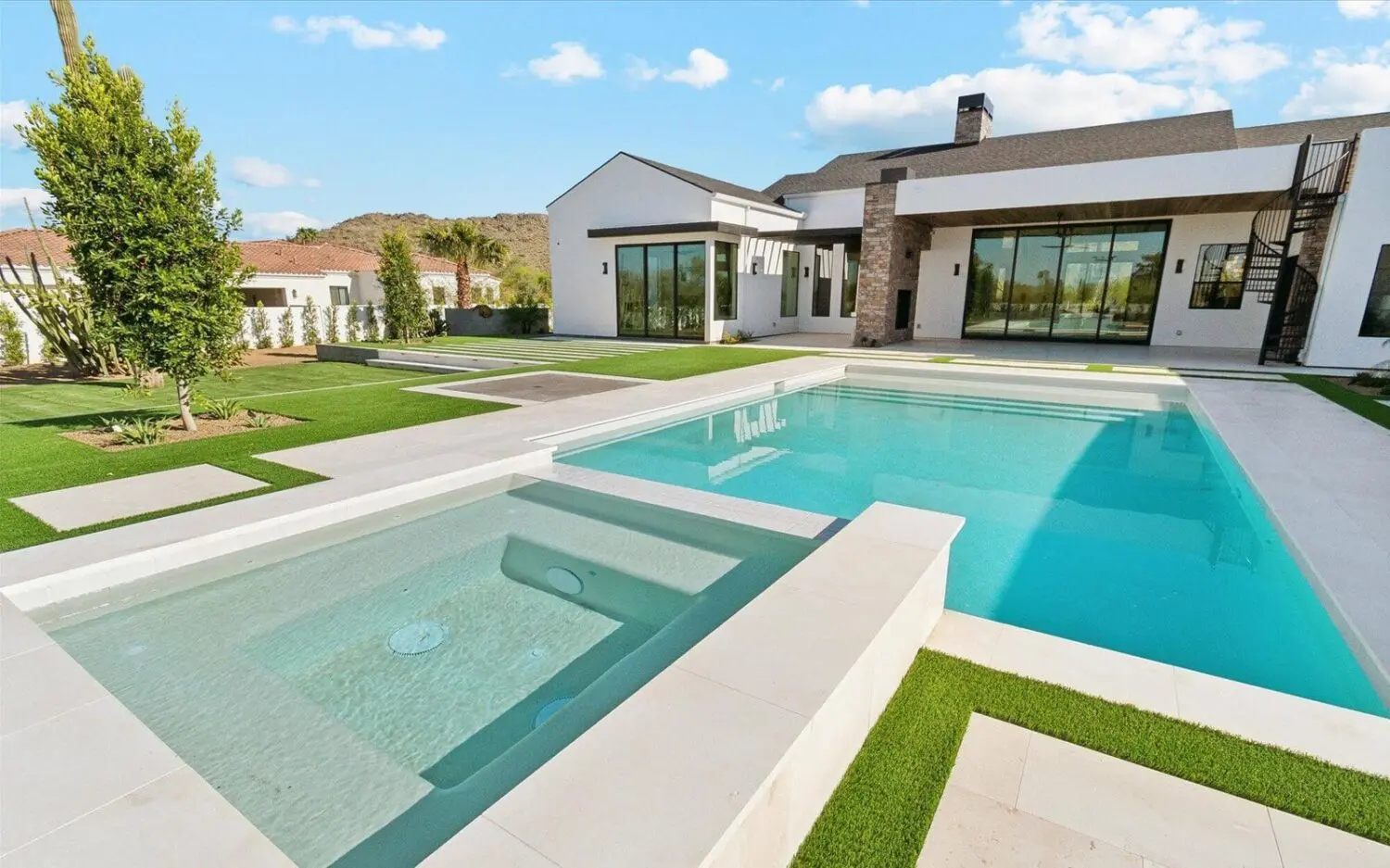 A backyard in Scottsdale boasts a putting green surrounded by desert landscaping. The area features small plants and palm-like trees along a stone-bordered edge, enhanced by artificial turf. A tall, brown stucco wall frames the scene with a clear blue sky in the background.