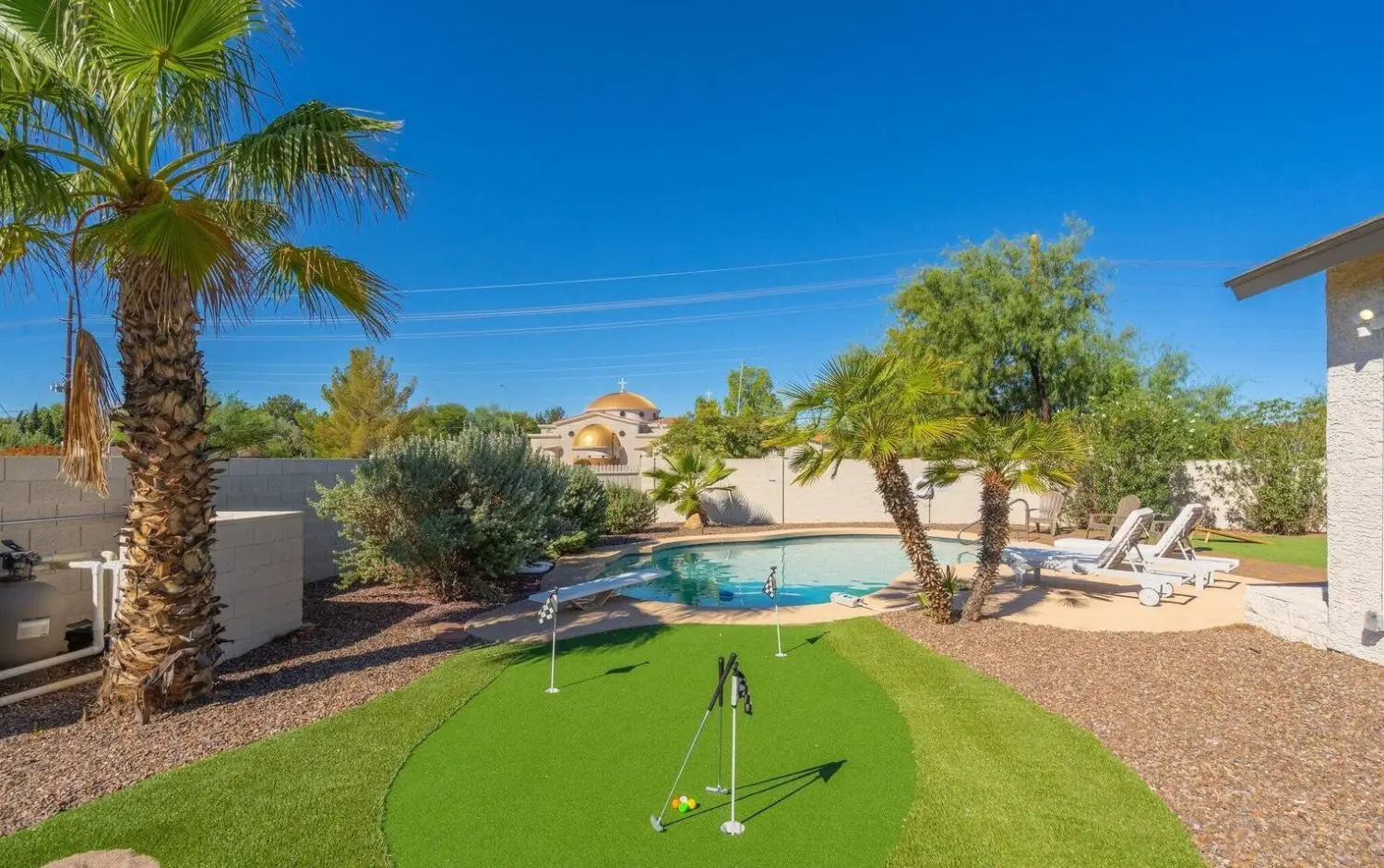 A backyard featuring a mini golf putting green with seamless artificial grass installation in the foreground, complemented by a swimming pool framed by lounge chairs, palm trees, and lush shrubs. Under a clear blue sky, a charming house stands proudly in the background.