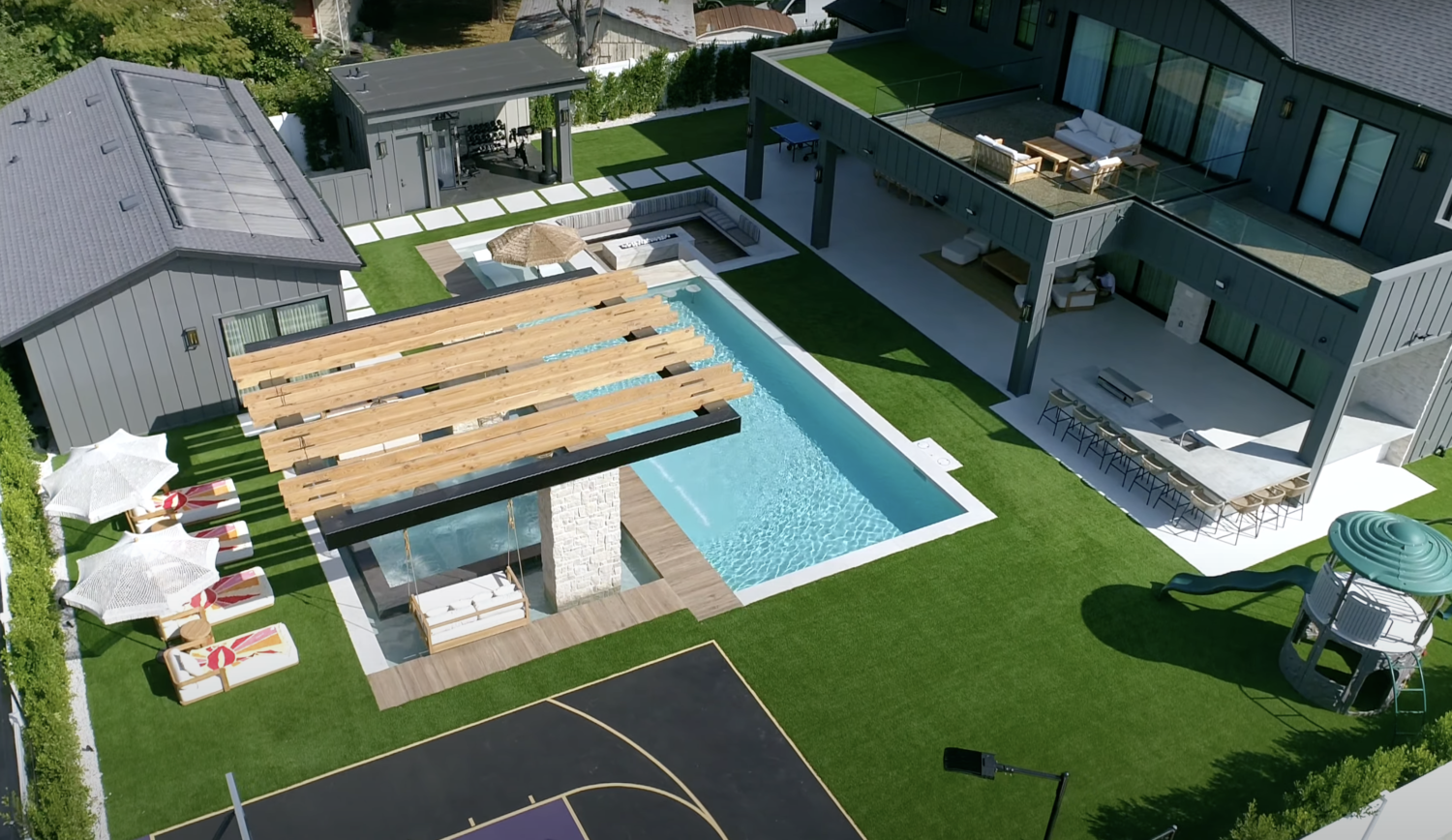 Aerial view of a modern backyard crafted by expert landscape designers in Scottsdale, featuring a swimming pool, poolside seating with umbrellas, a covered lounge area, and a basketball court adjacent to a two-story house with outdoor seating and a rooftop deck.