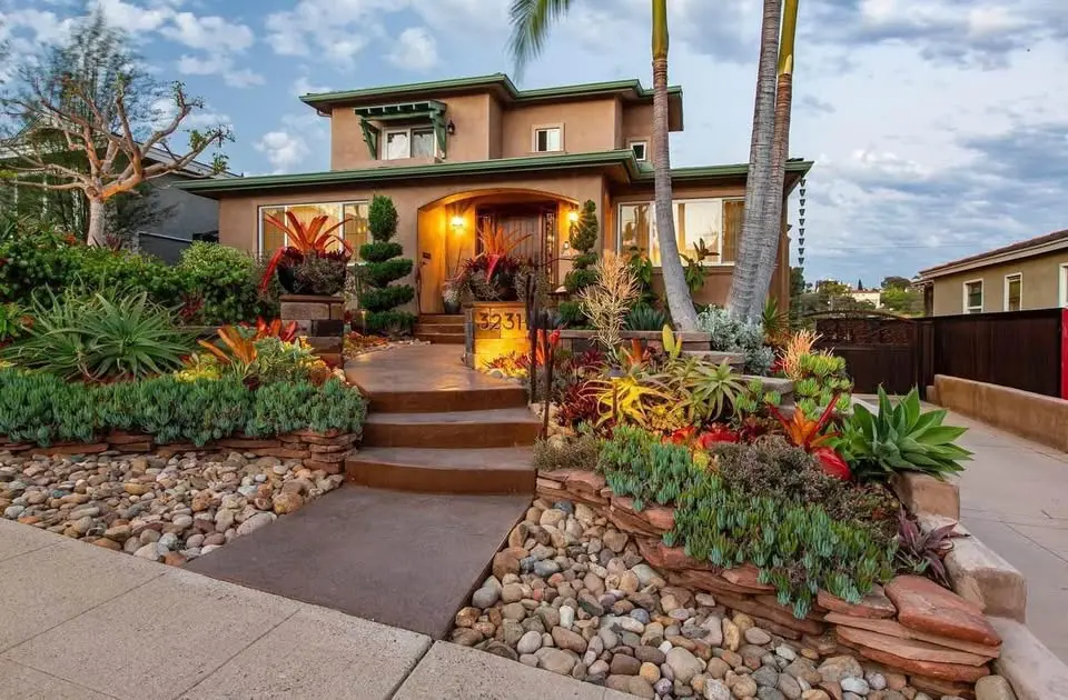 A two-story house with a green roof graces Scottsdale, surrounded by a landscaped front yard crafted by skilled landscape designers. Vibrant plants, rocks, and palm trees abound, while the pathway to the entrance is illuminated by warm outdoor lights.