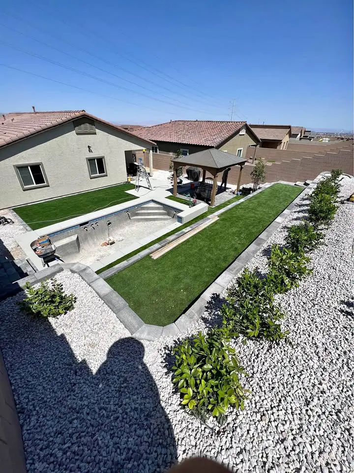 A backyard with a custom-designed rectangular in-ground pool under construction, surrounded by green artificial turf and white gravel. A wooden pergola stands to the right with several small bushes along the edge, all thoughtfully crafted by talented landscape designers. Houses are visible in the background.