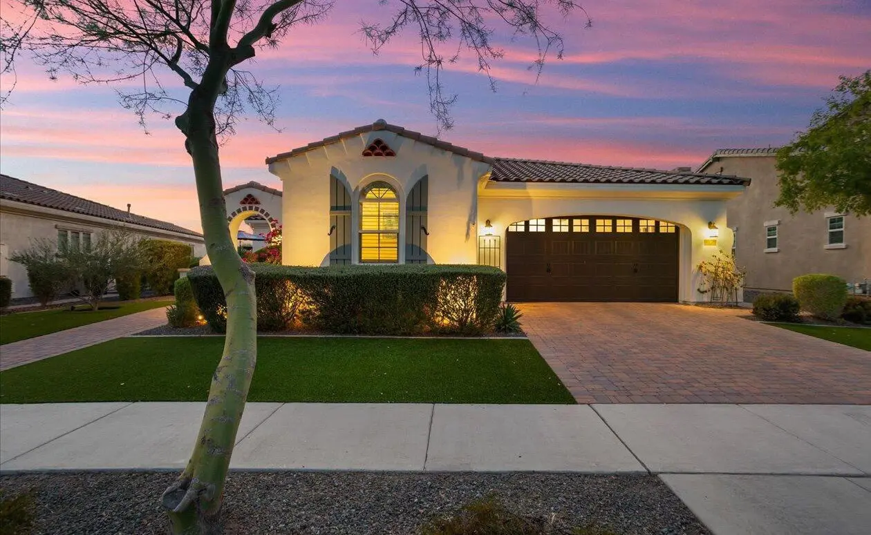 A single-story house in the Phoenix metro area boasts a stucco exterior and arched windows, framed by a neatly trimmed lawn and hedges. The driveway leads to a dark garage door, enhanced by subtle landscape lighting installation, while a pink and purple sunset paints the sky, silhouetting leafless branches.