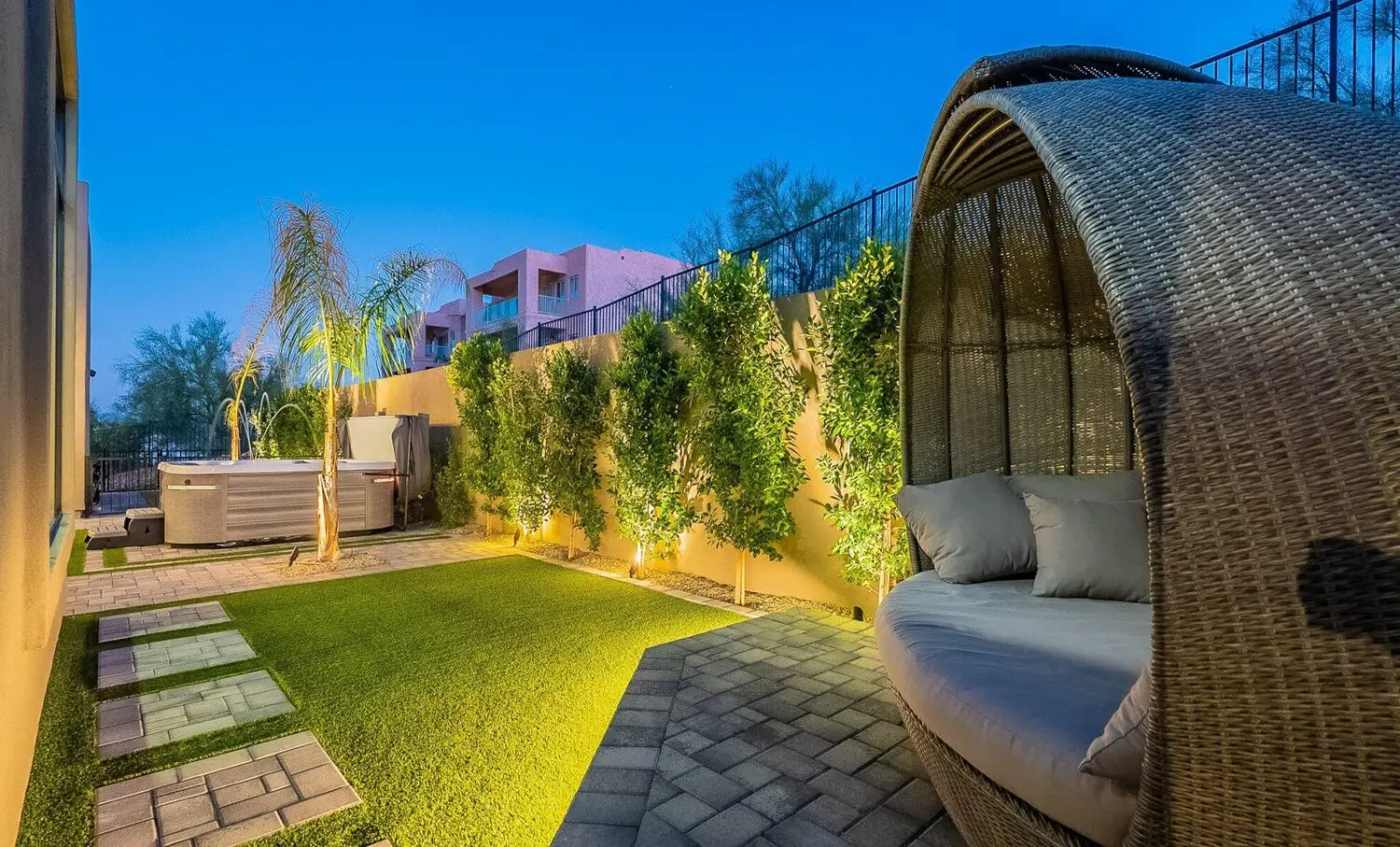 A cozy backyard in the Scottsdale area boasts a wicker daybed with cushions, a hot tub, and illuminated greenery thanks to expert landscape lighting installation. The ground is paved with stone tiles and artificial grass. The sky is a deep blue, hinting at early evening tranquility.