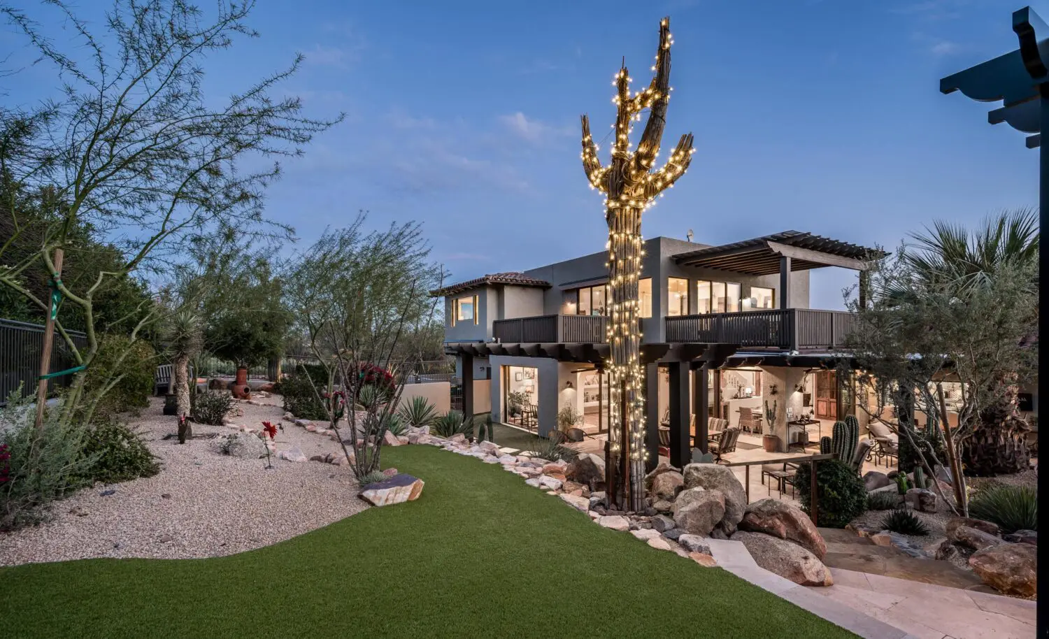 Luxurious two-story house with large windows, nestled in Scottsdale's desert landscaping. A tall cactus adorned with string lights stands prominently, enhanced by landscape lighting installation. The setting is during dusk, with a clear sky and ambient lights highlighting the exterior.