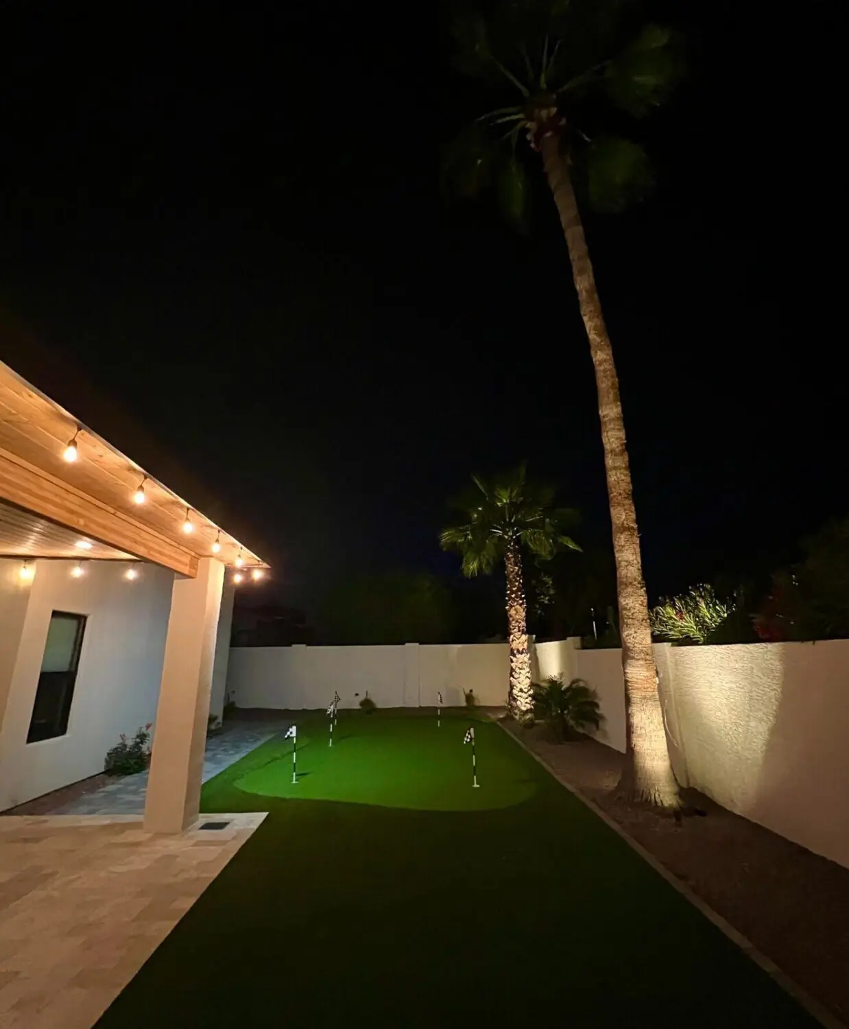 Nestled in the heart of the Phoenix metro area, this backyard at night boasts a cozy putting green with several golf holes. A tall palm tree and white fence create a serene border, while warm string lights illuminate the scene, showcasing expert landscape lighting installation under the wooden patio roof.