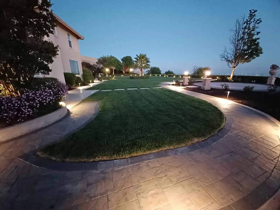 A curved pathway with embedded lights leads through a landscaped yard in the Scottsdale area, showcasing neatly trimmed grass. The path is bordered by flowerbeds, bushes, and a few trees. A house is visible on the left, and the deep blue sky suggests dusk.