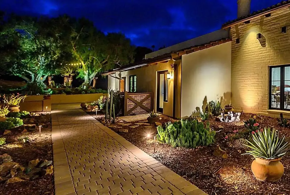 A well-lit path guides you to a house with a beige exterior and warm night lighting. The walkway, enhanced by expert landscape lighting installation, is adorned with small plants and lamps. Lush trees stand against the deep blue sky in the serene Phoenix metro area outdoor space.