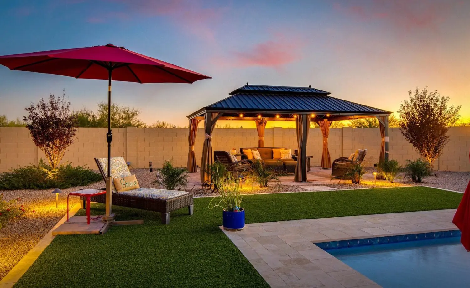 A cozy backyard scene at sunset in the Phoenix metro area features a pool in the foreground, a lounge chair with a red umbrella on the left, and a gazebo with cushioned seating beneath warm landscape lighting. Neatly landscaped greenery completes this Scottsdale haven.