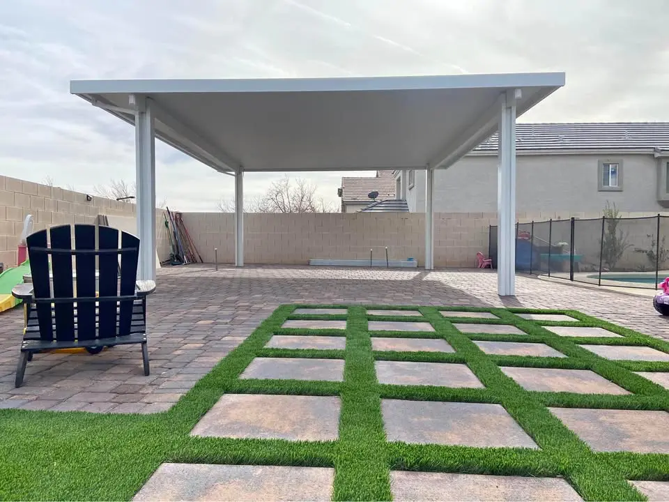 A single-story house with a beige stucco exterior in Scottsdale features a large covered patio and a well-manicured lawn, thanks to expert artificial turf installers. Tall trees and shrubs enhance the yard's charm under a clear blue sky.