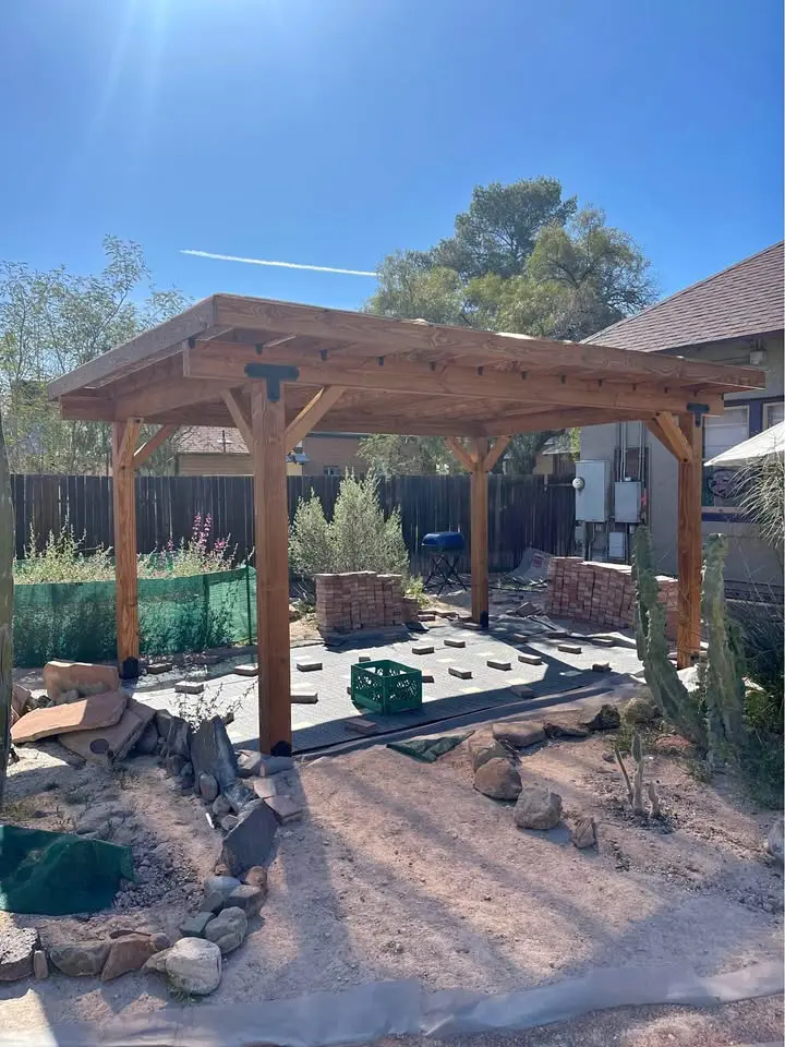 A wooden pergola graces a backyard, with a stone pathway meandering beneath it. Custom patio covers offer shade amidst lush green plants and rock landscaping. The sky remains clear and blue.