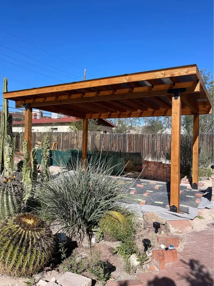 A custom pergola stands in a desert garden, surrounded by cacti and succulents, offering shade over the tiled patio. With a wooden fence and the clear Scottsdale AZ sky as its backdrop, this spot is perfect for enjoying serene desert beauty.