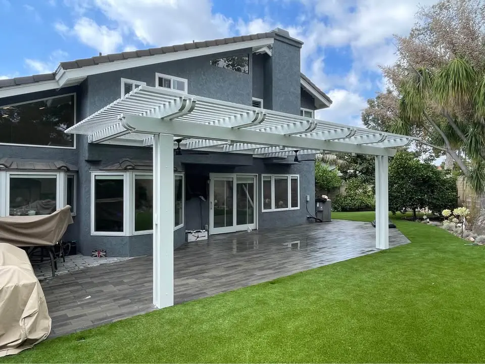 A modern gray two-story house with large windows and a white pergola over the patio features elegant gray tiles, offering a perfect spot to unwind while overlooking the lush green lawn. The sky is partly cloudy, and trees beautifully frame the backyard.