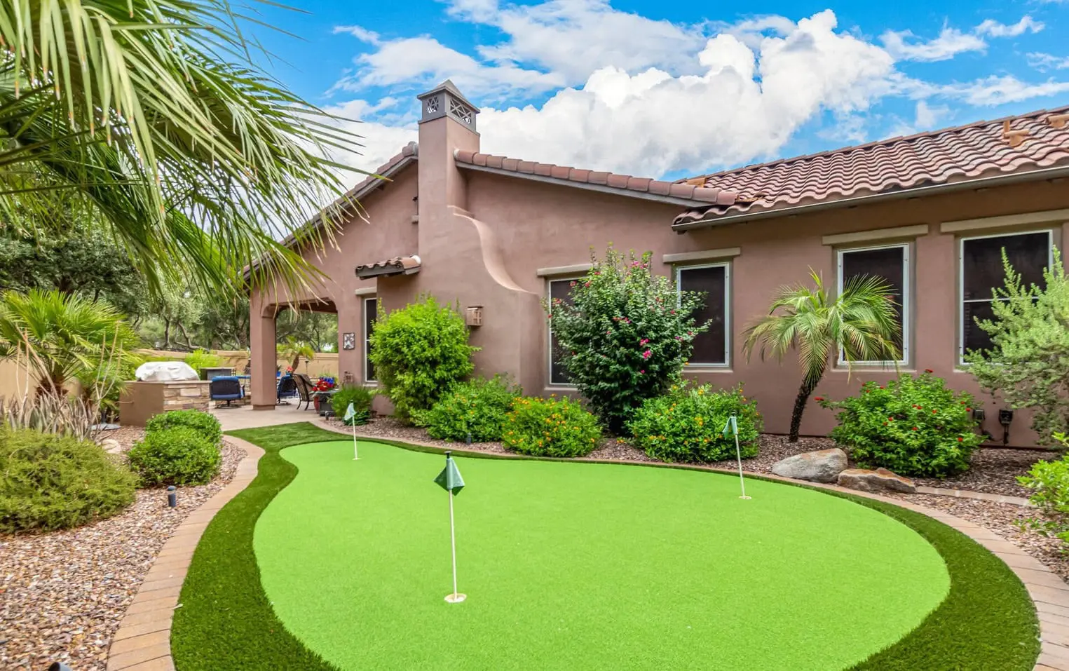 A Scottsdale backyard showcases a Mediterranean-style house with terracotta roof tiles. In the foreground, an artificial putting green is surrounded by lush landscaping with bushes, palm trees, and decorative rocks. The sky is partly cloudy, adding to the serene atmosphere.