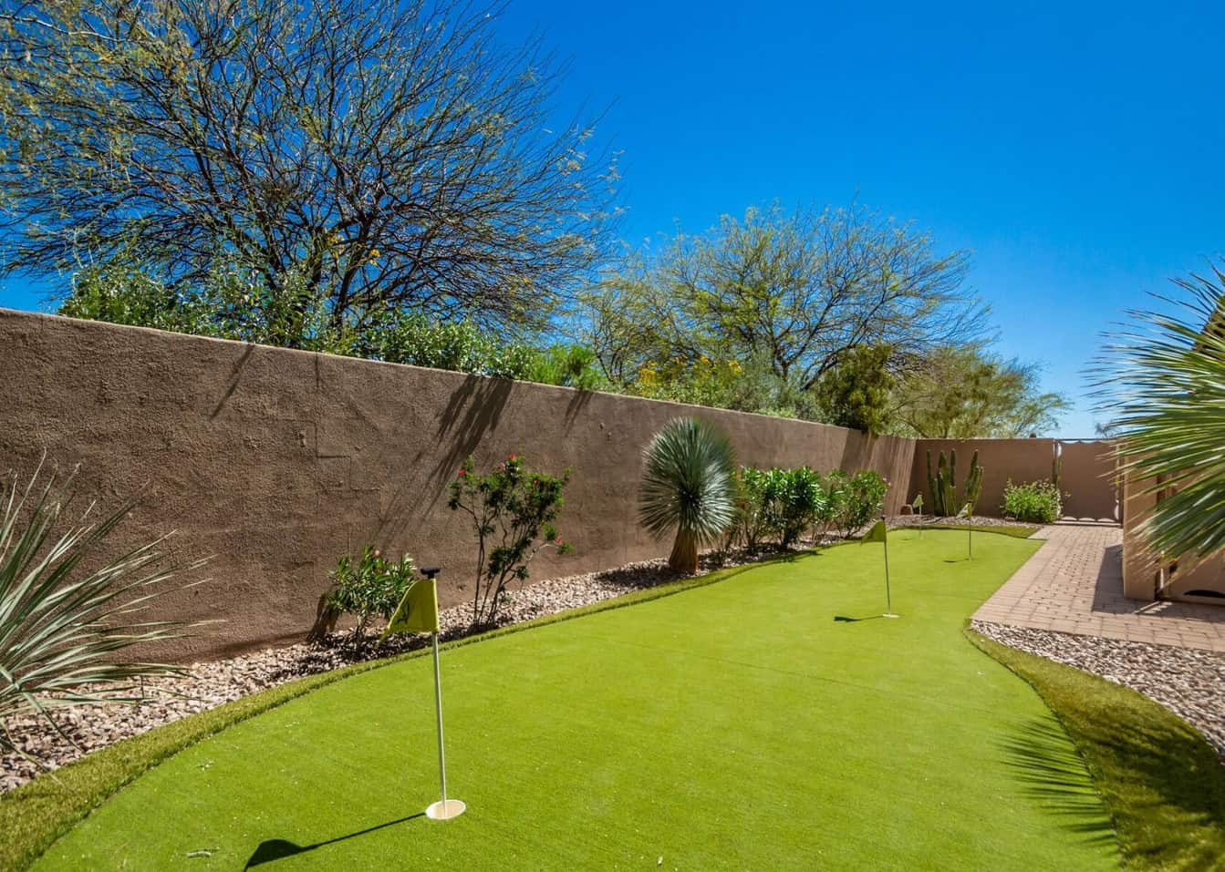 A Scottsdale backyard features a small putting green with several flags, surrounded by desert plants and trees. The lush artificial grass gives it an immaculate look. A stone pathway runs alongside a tall, brown wall under the clear blue sky.